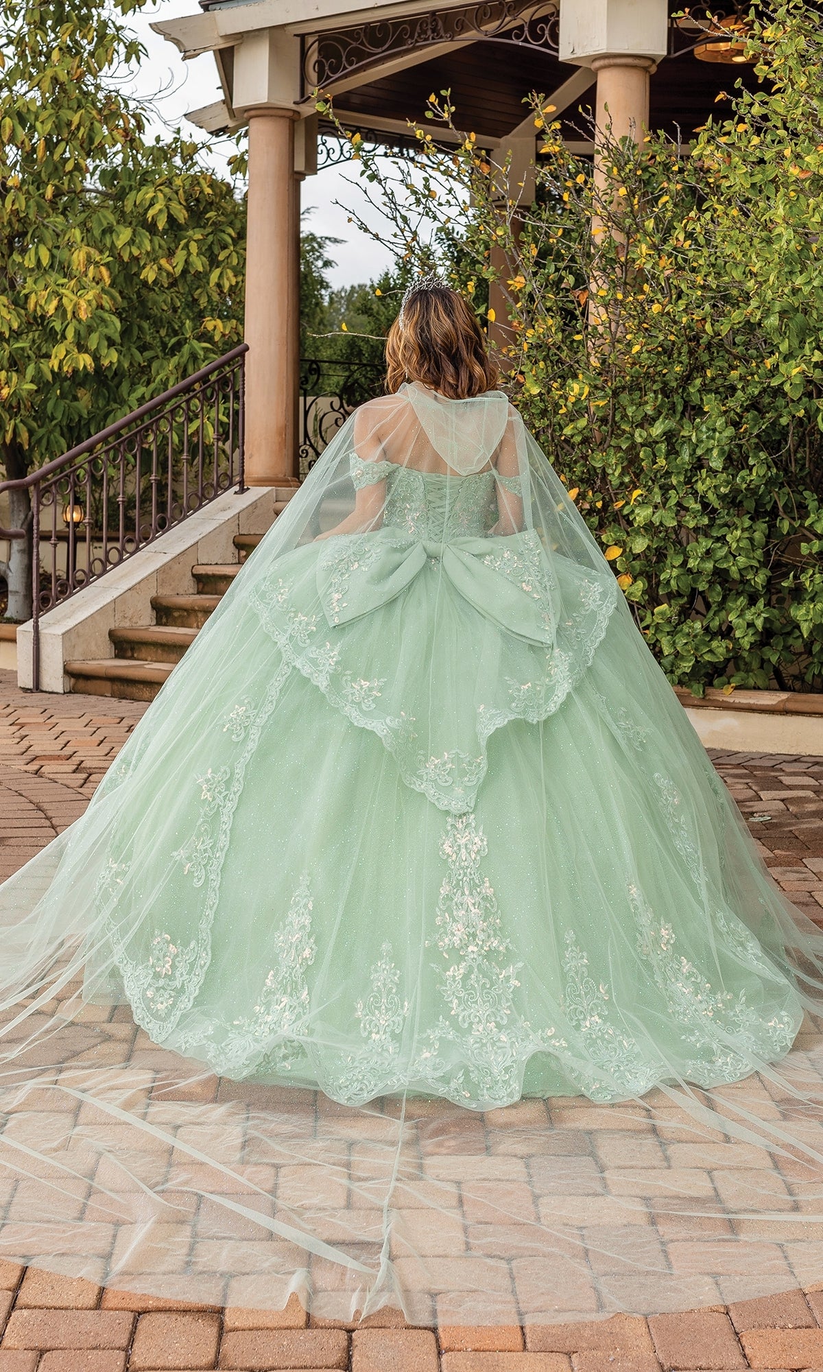 A person in the Quinceanera Dress 1843 By Dancing Queen, a light green a-line gown with lace details, stands outdoors facing away from the camera near a gazebo and greenery.